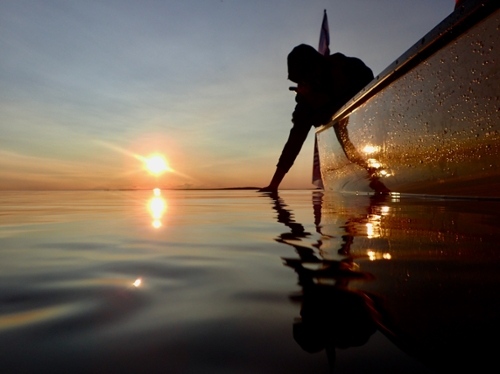 A bright sun hangs just above the waterline in a clear blue sky. The silhouette of a person reaches over the side of a canoe and has their hand in the water.   