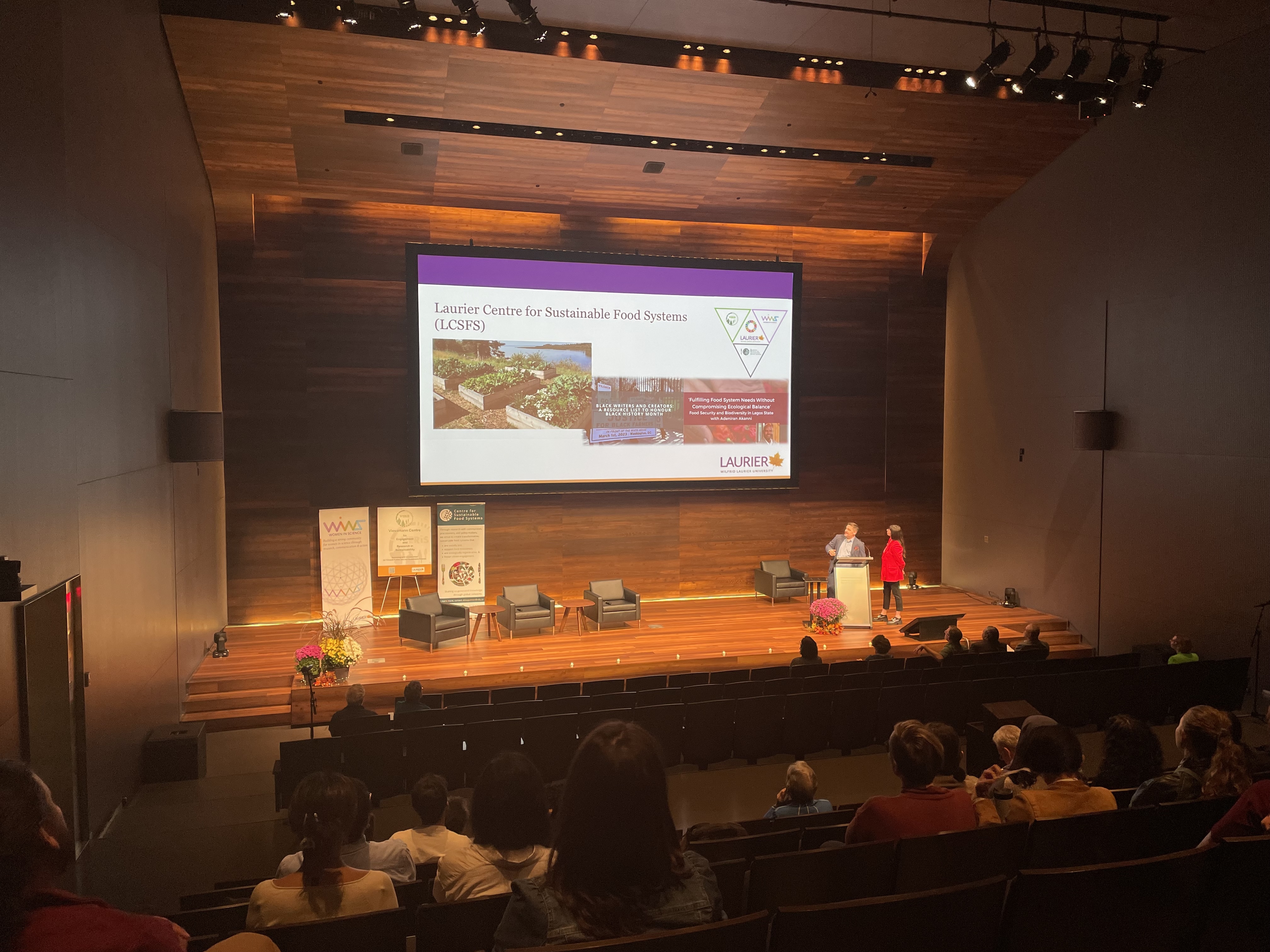 An audience faces a double screen presentation in a large auditorium