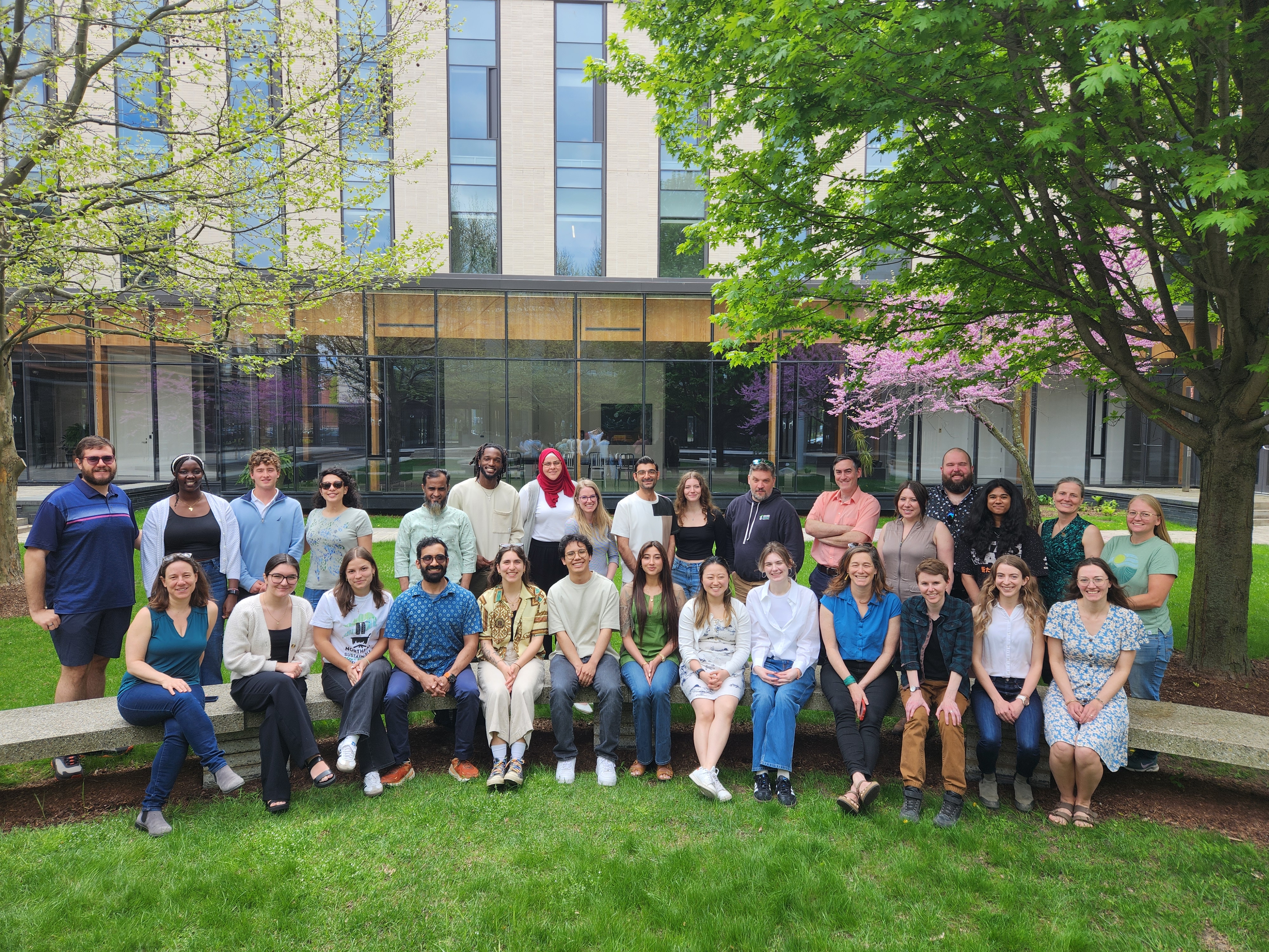 Group photo of a research lab, some people are sitting on a bench, with others standing behind them. They are outside it is spring, there are trees in the background