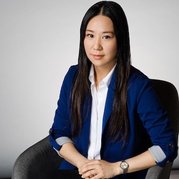 Woman in a blue blazer and white collared shirt, seated in a grey chair, with hands in her lap